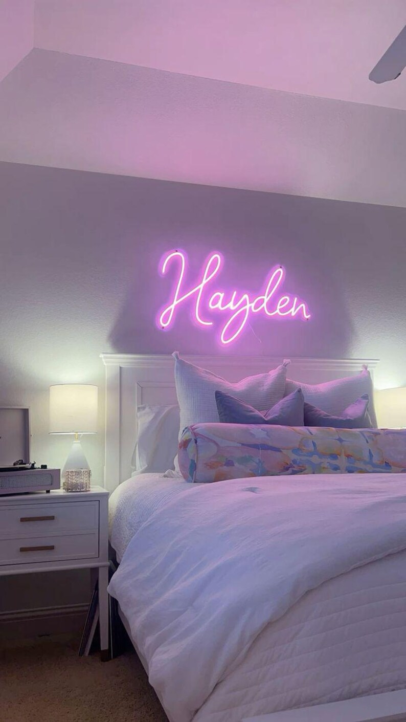 May include: A bedroom scene featuring a white bed with a white comforter and pillows. Above the bed, a neon sign spells out the name "Hayden" in a cursive font. A bedside table with a lamp and a record player is visible.