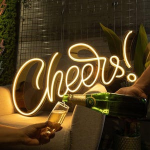 May include: A glowing yellow neon sign displaying the word Cheers in a cursive script font. The sign is positioned behind a clear glass flute being filled with a sparkling beverage from a green glass bottle, creating a festive atmosphere.