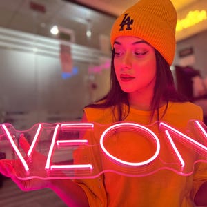 May include: A bright pink neon sign featuring the word NEON in capital letters, mounted on a clear acrylic backing. The sign emits a vibrant glow, illuminating the surrounding area with a distinct pink light, showcasing the linear tube design.
