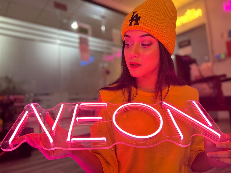 May include: A bright pink neon sign featuring the word NEON in capital letters, mounted on a clear acrylic backing. The sign emits a vibrant glow, illuminating the surrounding area with a distinct pink light, showcasing the linear tube design.