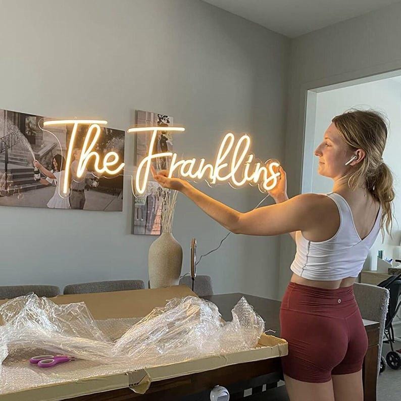May include: A custom neon sign featuring the text The Franklins in a warm, glowing script font. The sign is illuminated with a soft yellow light and is held up by a person. The lettering is connected in a continuous, flowing cursive style.