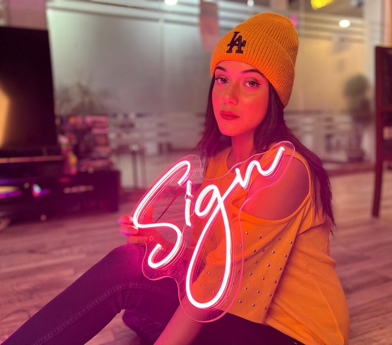 May include: A bright pink neon sign in cursive script that reads Sign, held by a person wearing a mustard yellow knit beanie with a dark blue LA logo and a matching mustard yellow cold-shoulder top with silver studs along the sleeves.