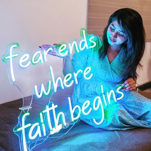 May include: A neon sign with the words "fear ends where faith begins" in white script. The sign has a clear backing and is illuminated with blue and green lights. The sign is held by a person wearing a blue dress.