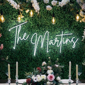 May include: A neon sign that reads "The Martings" in a cursive font, set against a backdrop of greenery and flowers. The scene is illuminated by hanging lights and candles, creating a romantic ambiance, perfect for a wedding or event.