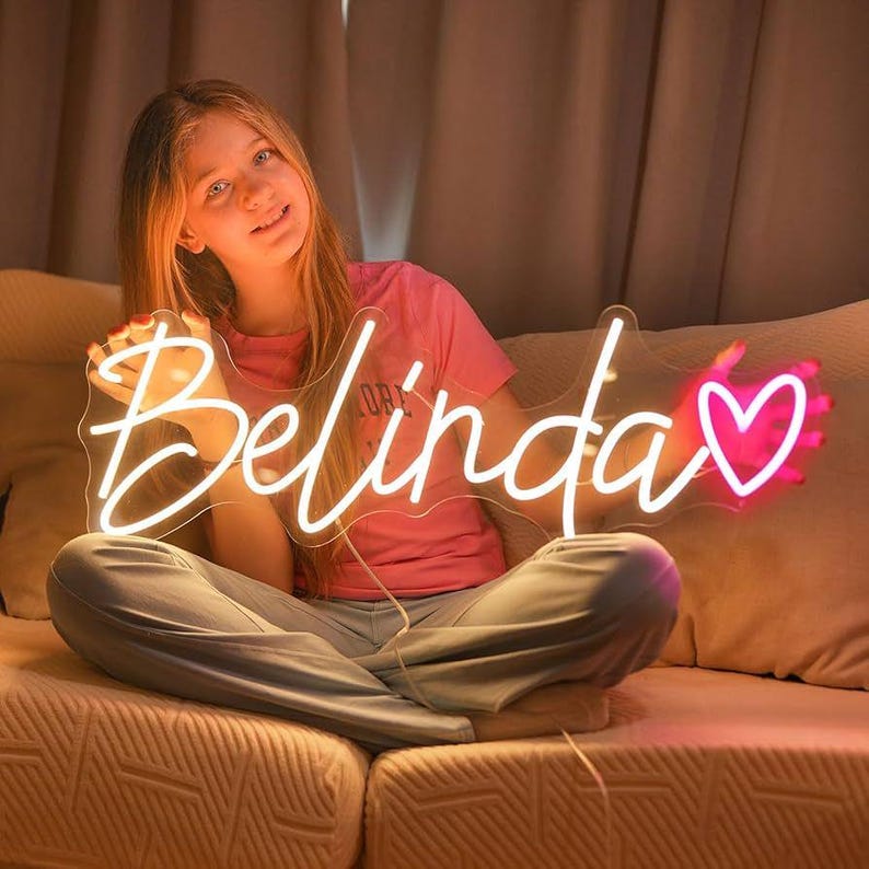 May include: A neon sign in the shape of the name "Belinda" with a pink heart. The sign is illuminated with a bright white light for the name and a pink light for the heart. The sign is held by a person.