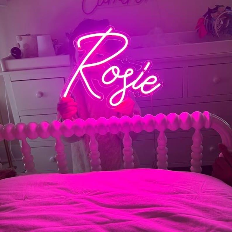 Puede incluir: Un letrero de ne&oacute;n rosa con el nombre "Rosie" en letras blancas. El letrero est&aacute; siendo sostenido por una persona frente a una cama blanca con una colcha rosa.