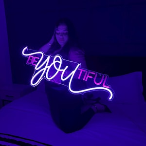 Könnte beinhalten: Ein Neon-Schild mit den Worten "Be You Tiful" in rosa und weißen Buchstaben. Das Schild wird von einer Person in einem dunklen Raum gehalten.
