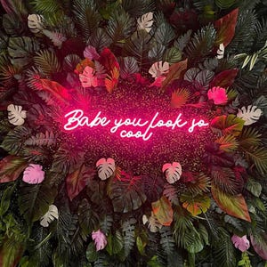 May include: A vibrant wall of lush greenery and tropical leaves surrounds a neon sign. The sign, in a bright pink hue, reads "Babe you look so cool" in a stylish script, creating a striking contrast against the natural backdrop.