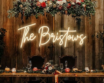 May include: A wedding reception scene featuring a wooden table set for two, adorned with floral arrangements, candles, and champagne glasses. Above, a neon sign reads "The Bristows" and a large floral arrangement hangs from the ceiling.