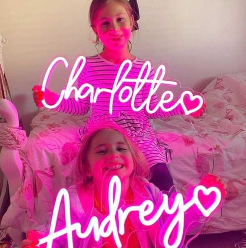 Puede incluir: Dos letreros de ne&oacute;n rosa con los nombres "Charlotte" y "Audrey" con un coraz&oacute;n al final de cada nombre. Los letreros est&aacute;n iluminados y los sostienen dos ni&ntilde;as.