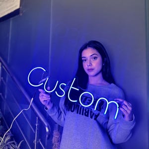 May include: A blue neon sign that spells out the word "Custom". The sign is illuminated with a bright, electric blue light. The sign is held in front of a person wearing a gray sweatshirt.
