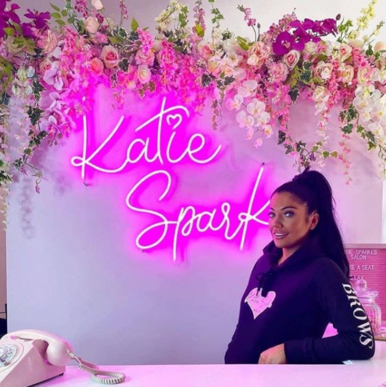 Puede incluir: Un letrero de ne&oacute;n rosa con el nombre "Katie Spark" en una fuente cursiva. El letrero est&aacute; colgado en una pared blanca decorada con flores rosas. Una mujer con cabello largo y oscuro est&aacute; de pie frente al letrero, usando una sudadera con capucha negra con un dise&ntilde;o de coraz&oacute;n rosa.