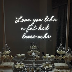 May include: A dessert table with various treats, including cupcakes, cake pops, and cookies. The phrase "Love you like a fat kid loves cake" is written in neon lights above the table. The desserts are arranged on glass platters and trays.