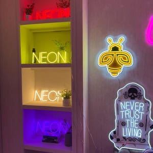 May include: A white shelf unit with four shelves displaying neon signs. The top shelf has a red and pink neon sign that says "NEON". The second shelf has a yellow neon sign that says "NEON". The third shelf has a yellow neon sign that says "NEON". The bottom shelf has a purple neon sign that says "NEON". There is also a yellow neon bee sign and a white neon sign that says "NEVER TRUST THE LIVING" on the wall behind the shelf unit.