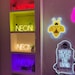 May include: A white shelf unit with four shelves displaying neon signs. The top shelf has a red and pink neon sign that says "NEON". The second shelf has a yellow neon sign that says "NEON". The third shelf has a yellow neon sign that says "NEON". The bottom shelf has a purple neon sign that says "NEON". There is also a yellow neon bee sign and a white neon sign that says "NEVER TRUST THE LIVING" on the wall behind the shelf unit.