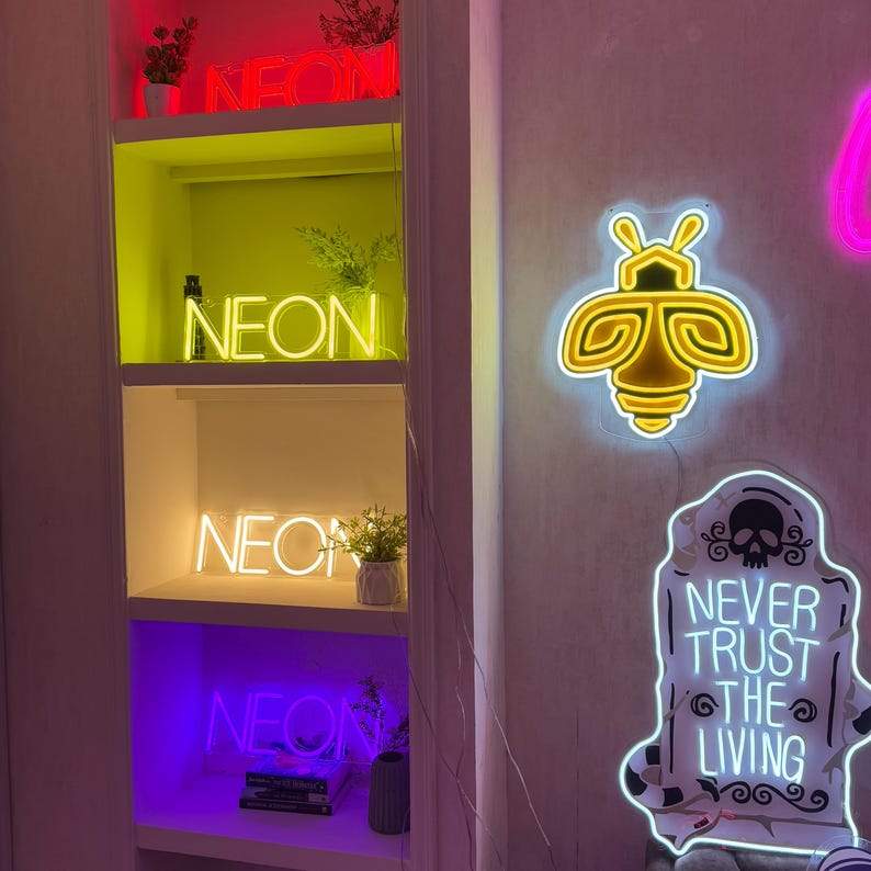 May include: A white shelf unit with four shelves displaying neon signs. The top shelf has a red and pink neon sign that says "NEON". The second shelf has a yellow neon sign that says "NEON". The third shelf has a yellow neon sign that says "NEON". The bottom shelf has a purple neon sign that says "NEON". There is also a yellow neon bee sign and a white neon sign that says "NEVER TRUST THE LIVING" on the wall behind the shelf unit.