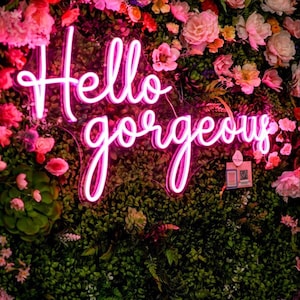 May include: A neon sign with the words "Hello gorgeous" in a cursive font, illuminated in pink. The sign is set against a backdrop of lush greenery and a variety of colorful flowers, including pink and orange roses.