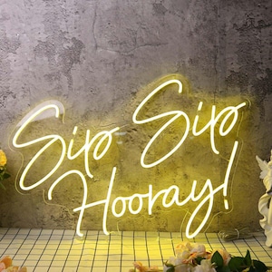 May include: A neon sign with the words "Sip Sip Hooray!" in a cursive font. The sign glows with a warm yellow light against a textured gray background. The sign is a decorative piece for a bar or event.