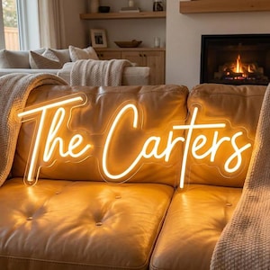 May include: A custom neon sign featuring the text The Carters in a warm, glowing yellow script font. The sign is mounted on a clear acrylic backing and is positioned against a tan leather sofa, emitting a bright light that illuminates the surface.