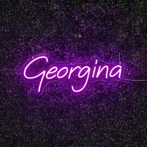 May include: A neon sign in a cursive font spelling the name "Georgina" in bright purple. The sign is mounted on a dark green textured background, likely a wall of foliage. The neon light is a vibrant, eye-catching color.