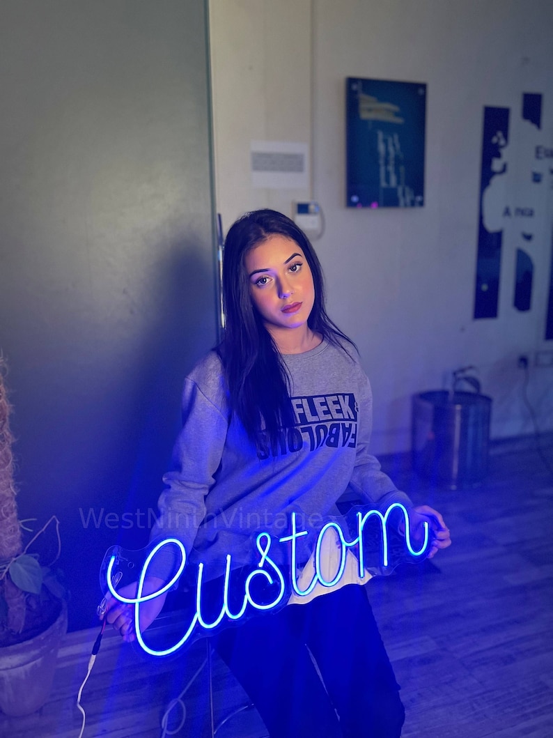 May include: A woman is holding a blue neon sign that reads "Custom". The sign is in a cursive font. The woman is wearing a grey sweatshirt with the words "Fabulous Fleek" printed on it.