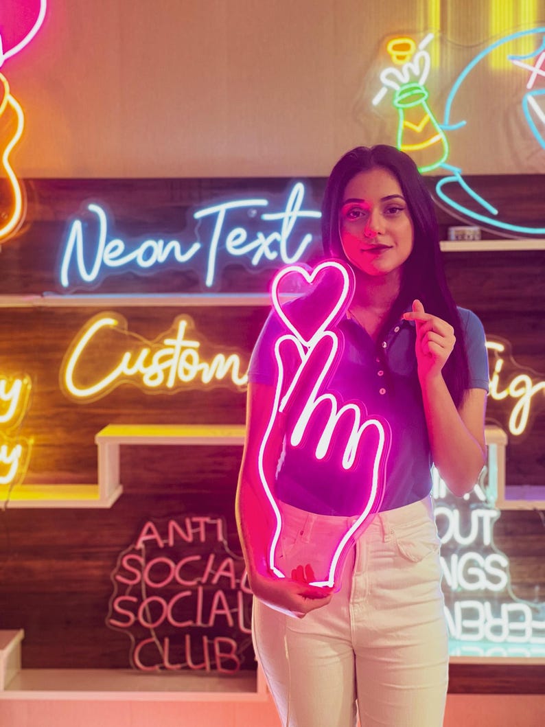 May include: A pink neon sign shaped like a hand with a heart on the index finger. The sign is held by a person in front of a wall with other neon signs, including one that says "Neon Text Custom".