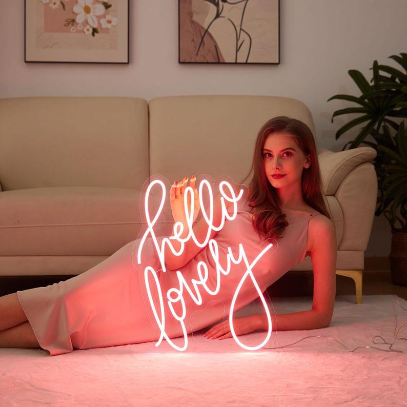 May include: A bright white neon sign featuring the cursive text hello lovely, illuminated with a glowing light. The sign is constructed with a clear acrylic backing and is powered by a thin electrical cord extending from the bottom edge.