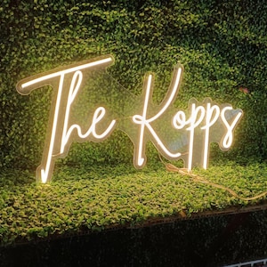 May include: A neon sign with the words "The Kopps" in a cursive font. The sign is illuminated with a warm, white light and is set against a backdrop of green foliage. The sign is likely used for home decor or event decoration.