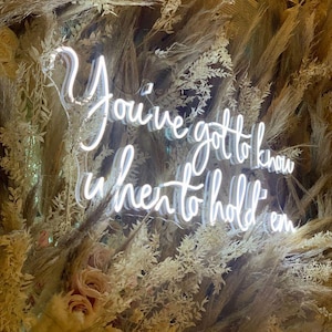 May include: A neon sign with the words "You've got to know when to hold em" in a cursive font. The sign is surrounded by dried plants and flowers, creating a natural and rustic aesthetic.