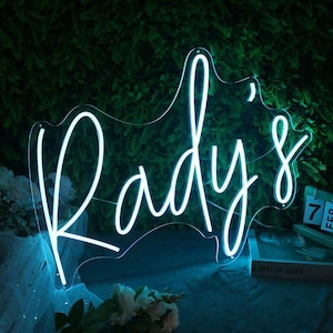 May include: A neon sign in the shape of a silhouette, with the word "Rady's" in a cursive font. The sign glows with a bright, cool blue light against a backdrop of green foliage. A calendar shows the date.