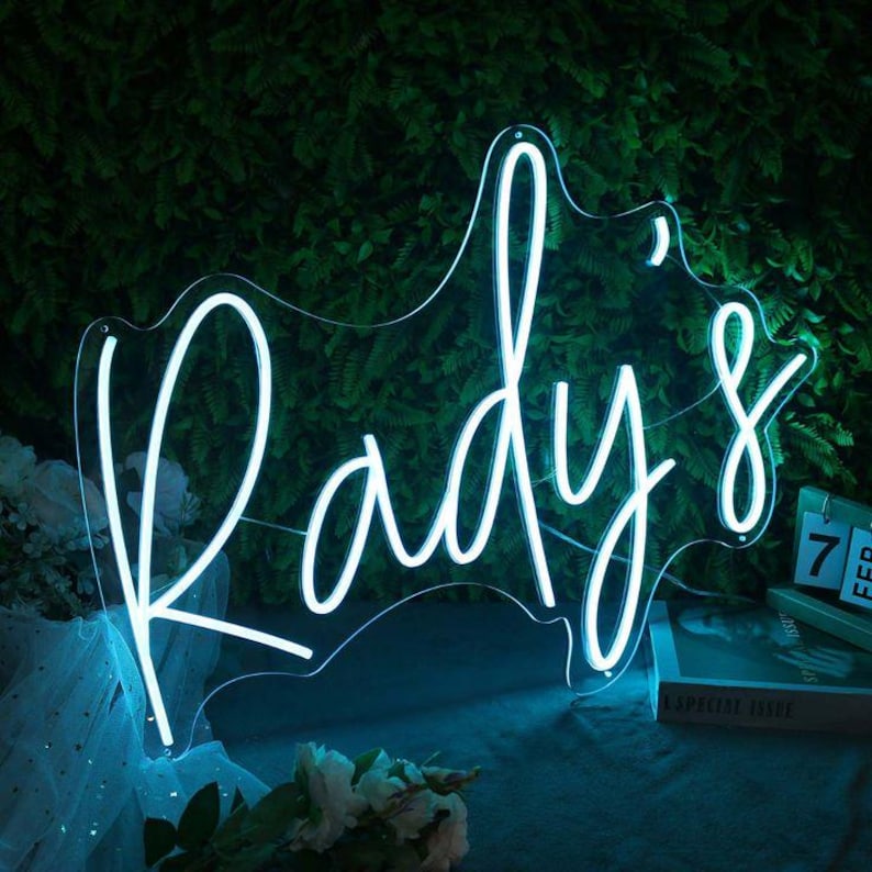 May include: A neon sign in the shape of a silhouette, with the word "Rady's" in a cursive font. The sign glows with a bright, cool blue light against a backdrop of green foliage. A calendar shows the date.