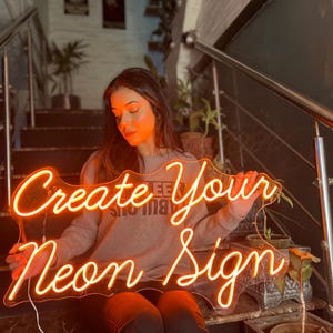 May include: A custom neon sign featuring the orange glowing text Create Your Neon Sign in a cursive script font. The sign is mounted on a clear acrylic backing and emits a warm light, displaying the words clearly against the dark background.