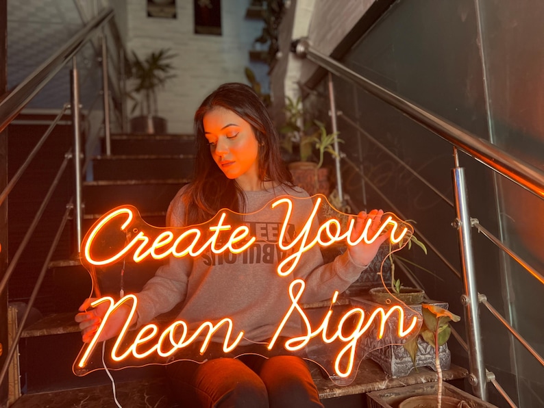 May include: A custom neon sign featuring the orange glowing text Create Your Neon Sign in a cursive script font. The sign is mounted on a clear acrylic backing and emits a warm light, displaying the words clearly against the dark background.