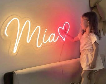 May include: A wall-mounted neon sign displaying the name Mia in white cursive script followed by a red neon heart outline. The sign is mounted on a plain wall above a light-colored upholstered headboard, emitting a soft glow in the room.