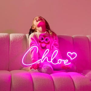 以下が含まれることがあります： 「Chloe」という名前とハートがついたピンクのネオンサイン。サインは点灯しており、白いソファの上に置かれています。若い女の子がソファに座ってサインを見上げています。