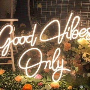 May include: A bright white neon sign featuring the text Good Vibes Only in a cursive script font. The sign is mounted on a clear acrylic backing and is positioned in front of a display of artificial flowers and greenery with a white stool nearby.