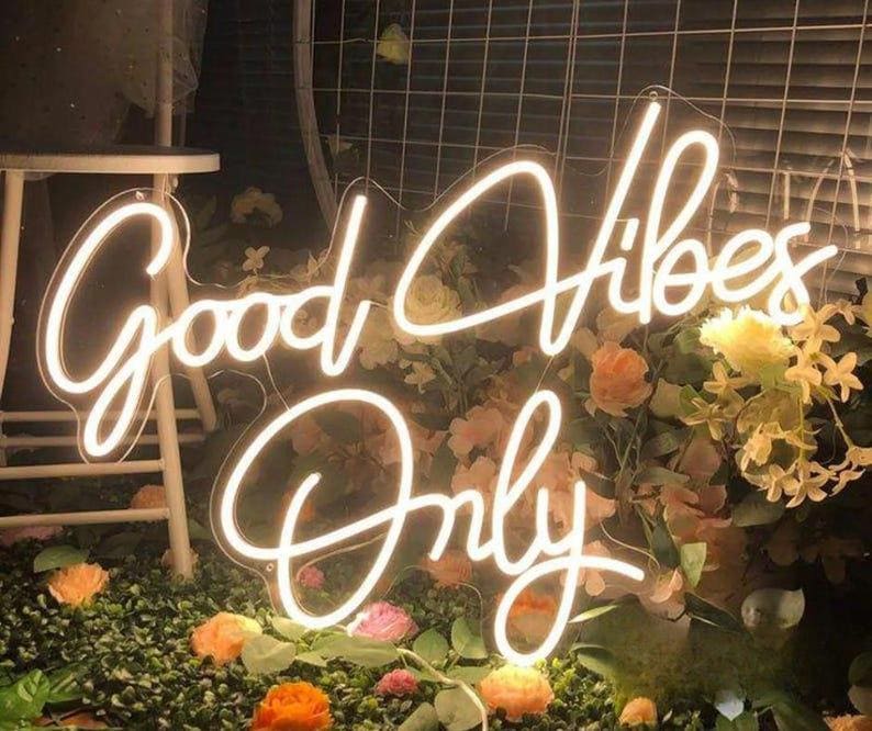 May include: A bright white neon sign featuring the text Good Vibes Only in a cursive script font. The sign is mounted on a clear acrylic backing and is positioned in front of a display of artificial flowers and greenery with a white stool nearby.