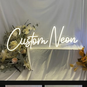 May include: A custom neon sign featuring the words Custom Neon in a white glowing script font. The sign is mounted on a clear acrylic backing and is displayed alongside floral arrangements with white and yellow flowers on a draped white fabric.