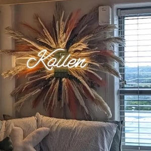 May include: A wall-mounted decorative arrangement featuring a sunburst of dried pampas grass in shades of cream, tan, and muted pink. Centered within the foliage is a glowing neon sign displaying the name Kailen in a white cursive script font.