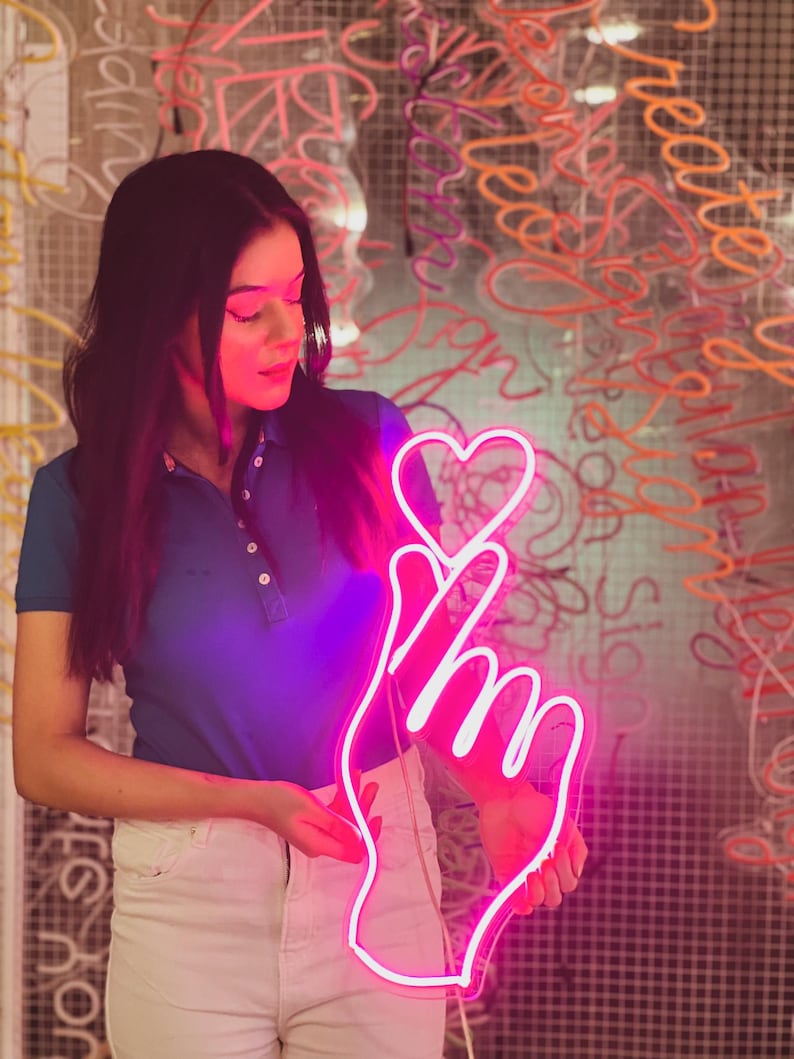 May include: A pink neon sign shaped like a hand making a pinky promise gesture with a heart on the index finger.