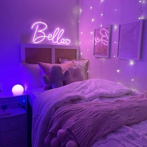 May include: A bedroom bathed in purple light features a bed with a pink knitted throw and pillows. A neon sign that says "Bellas" hangs above the bed. String lights and framed art decorate the walls, creating a cozy atmosphere.