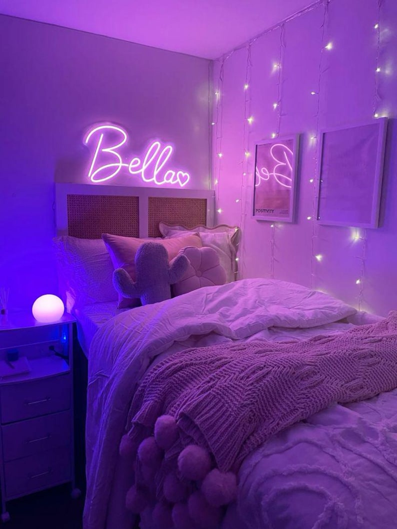 May include: A bedroom bathed in purple light features a bed with a pink knitted throw and pillows. A neon sign that says "Bellas" hangs above the bed. String lights and framed art decorate the walls, creating a cozy atmosphere.