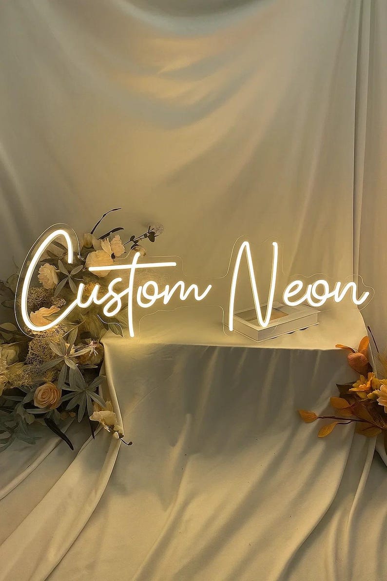 May include: A custom neon sign featuring the words Custom Neon in a white cursive script font. The sign is mounted on a clear acrylic backing and is illuminated with a bright white glow, positioned alongside decorative artificial flowers.