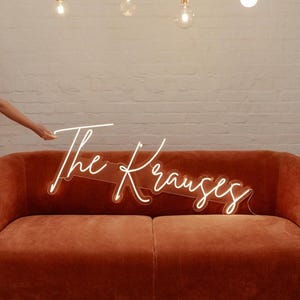 May include: A custom neon sign featuring the text The Krauses in a white cursive script font, mounted on a clear acrylic backing. The sign is positioned above a solid burnt orange velvet sofa, creating a bright and illuminated display piece.