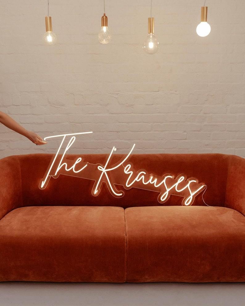 May include: A custom neon sign featuring the text The Krauses in a white cursive script font, mounted on a clear acrylic backing. The sign is positioned above a solid burnt orange velvet sofa, creating a bright and illuminated display piece.