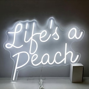 May include: A white neon sign with the phrase "Life's a Peach" in a cursive font. The sign is mounted on a clear backing and includes a power cord and a small white box.