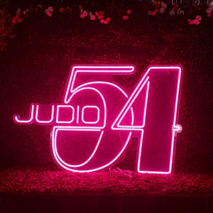May include: A neon sign displaying the text "JUDIO 54" in a vibrant pink hue. The numbers and letters are outlined with bright neon lights, creating a striking visual against a dark background. The sign is likely for a business or event.