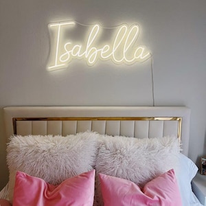 May include: A bedroom scene featuring a glowing neon sign that spells "Isabella" in a cursive font. Below the sign, a bed is adorned with fluffy white pillows and vibrant pink velvet pillows, creating a cozy and stylish aesthetic.