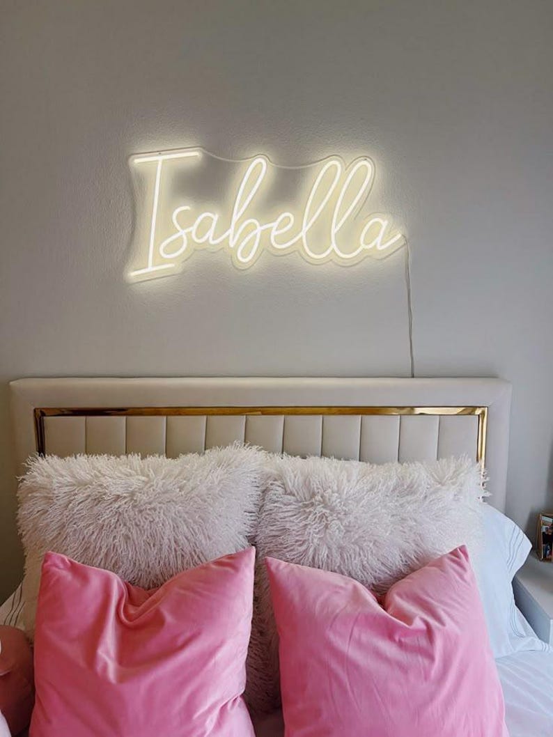 May include: A bedroom scene featuring a glowing neon sign that spells "Isabella" in a cursive font. Below the sign, a bed is adorned with fluffy white pillows and vibrant pink velvet pillows, creating a cozy and stylish aesthetic.