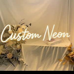 May include: A white neon sign featuring the script text Custom Neon, illuminated with a bright glow. The sign is positioned near a floral arrangement with light-colored flowers and foliage, resting on a draped fabric surface with a book.